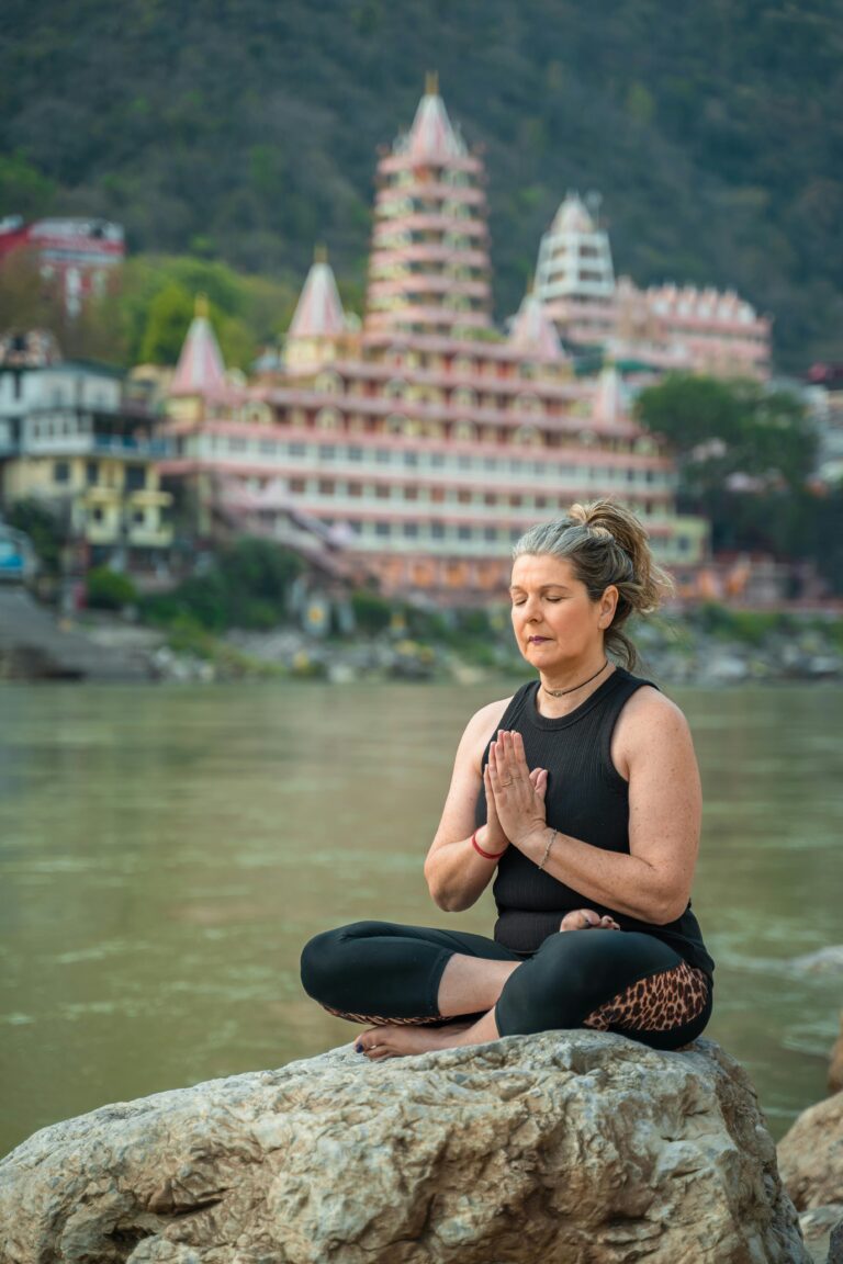 Woman meditating in lotus position by River Ganges with Rishikesh temple backdrop, symbolizing peace and spirituality.